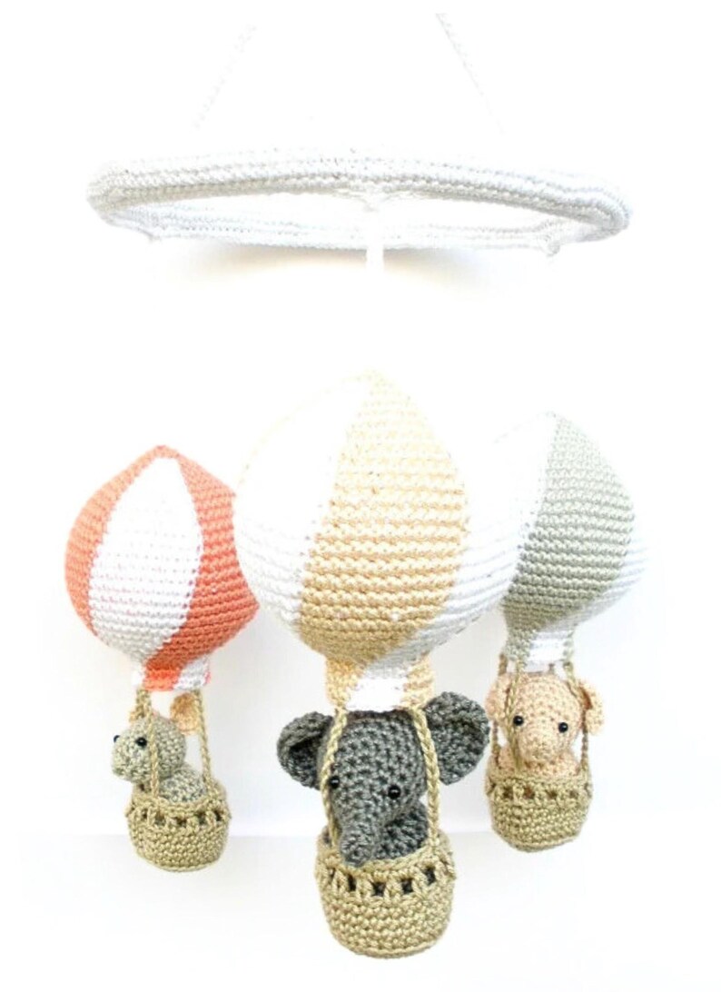 May include: A crocheted baby mobile with three hot air balloons. Each balloon is striped with white and a different color: peach, beige, and gray. Each balloon has a crocheted elephant in a basket hanging from it.