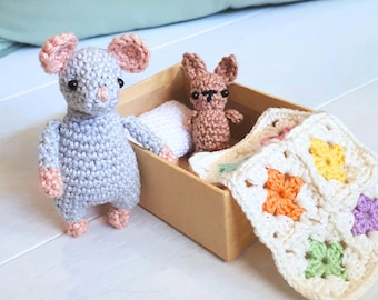 May include: A grey crocheted mouse, a brown crocheted bunny, and a white crocheted blanket with colourful squares are in a cardboard box.