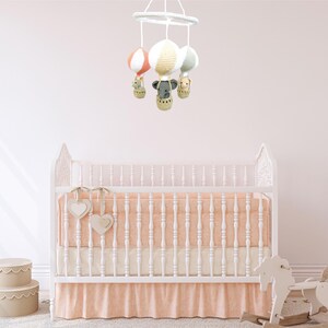 May include: A white crib with a peach-colored skirt and a white mobile hanging above it. The mobile has four hot air balloons with animal figures inside. The crib is in a nursery with a white rug and a rocking horse.