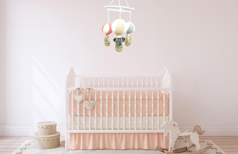 May include: A white crib with a peach-colored skirt and a white mobile hanging above it. The mobile has four hot air balloons with animal figures inside. The crib is in a nursery with a white rug and a rocking horse.
