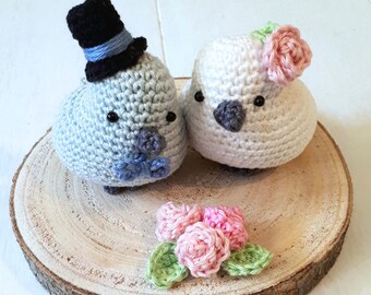May include: Two crocheted birds, one in light blue and one in white, wearing a top hat and a flower crown, respectively. They are standing on a wooden slice with a bouquet of pink crocheted flowers.