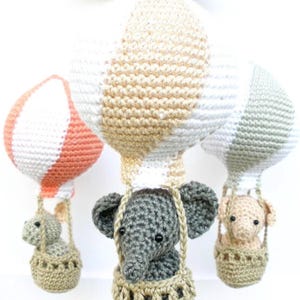 May include: Three crocheted hot air balloons with elephants in baskets. The balloons are white, peach, and gray with beige stripes. The elephants are gray, peach, and beige.