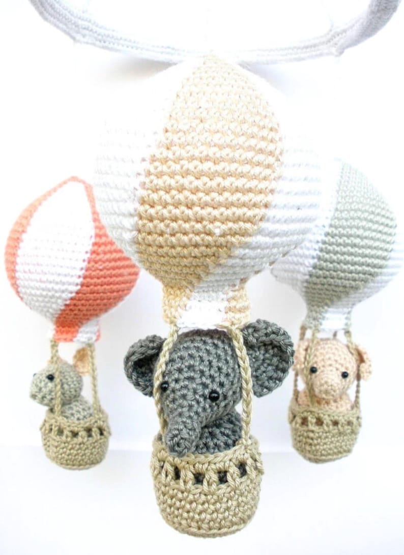 May include: Three crocheted hot air balloons with elephants in baskets. The balloons are white, peach, and gray with beige stripes. The elephants are gray, peach, and beige.