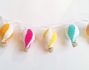 May include: A garland of five crocheted hot air balloons in yellow, white, pink, blue, and orange. The balloons are strung together with white yarn.