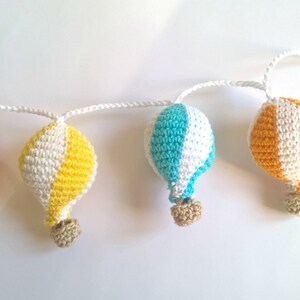 May include: A garland of five crocheted hot air balloons in red, white, yellow, and blue. The balloons are connected by a white string.