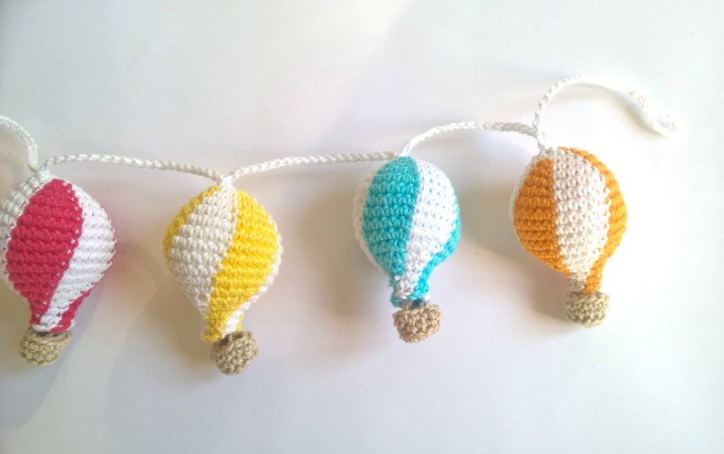 May include: A garland of five crocheted hot air balloons in red, white, yellow, and blue. The balloons are connected by a white string.