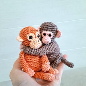 May include: Two crocheted monkey dolls. One is orange, the other is brown. The brown monkey is hugging the orange monkey. Both have black eyes and pink accents on their ears and faces. The dolls are handmade.