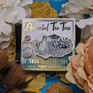 Puede incluir: Dos pines de esmalte con un leopardo de las nieves en una tetera y un leopardo de las nieves en una taza. Los pines son dorados con detalles azules y blancos. El texto "Elemental Tea Time" y "Snow Leopard Tea" está en la tarjeta.