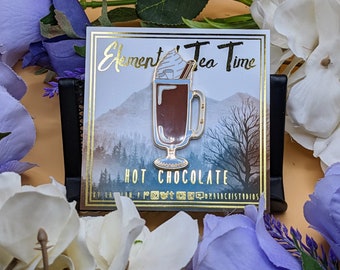 May include: A pin featuring a hot chocolate design with whipped cream and a cinnamon stick. The pin is on a card with the text "Element Tea Time" and "Hot Chocolate". The background shows a mountain scene.