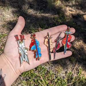 May include: Five enamel pins featuring swords with floral designs. The swords are decorated with red, white, blue, and orange flowers. The pins are gold-toned.