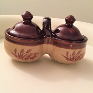 May include: A vintage ceramic condiment server with two connected compartments and matching lids. The server is tan with brown accents and wheat stalk designs. A handle connects the two sections.
