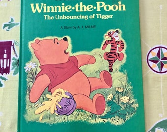 Puede incluir: Una cubierta de libro verde con el título "Winnie de Pooh El rebote de Tigger" en letras amarillas. El libro está ilustrado con Winnie de Pooh sosteniendo un tarro de miel y Tigger de pie detrás de él.