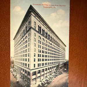 May include: Vintage postcard featuring the Wanamaker Building in Philadelphia, Pennsylvania, as seen from City Hall. The postcard shows a large, multi-story building with numerous windows and architectural details, with cars parked on the street.
