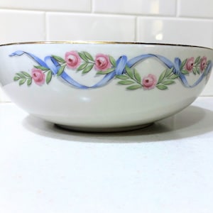 May include: A white ceramic bowl featuring a decorative band around the upper exterior. The design consists of pink roses, green leaves, and light blue ribbons arranged in a repeating garland pattern. The rim of the bowl is finished with a gold trim.