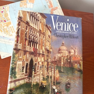 May include: A hardcover book titled Venice: The Biography of a City by Christopher Hibbert, featuring a painting of Venetian canals and architecture on the cover, positioned next to a partially visible map of the city.