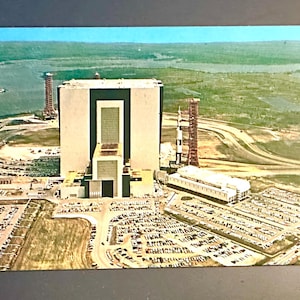 Op de afbeelding: Een luchtfoto van een groot, wit gebouw met een zwart dak. Het gebouw is omgeven door een parkeerplaats en een grasveld. Er is een hoge, witte toren op de achtergrond.
