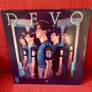May include: Album cover for Devo's "New Traditionalists." The album art features five figures in blue shirts and gray pants, with the word "DEVO" in white at the top. Each figure has a patch on the sleeve with a portrait.