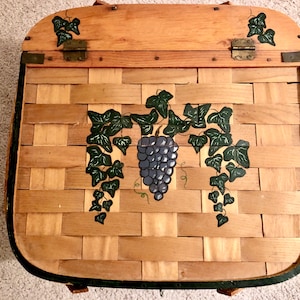 May include: A square woven wood basket with a hinged lid featuring a hand-painted design of a cluster of purple grapes and green ivy leaves. The basket has a natural light brown wood finish, dark green painted edges, and metal hinges.