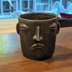 May include: A dark gray ceramic planter with a face design. The planter has a cylindrical shape with a wide opening and two small handles. The face features closed eyes, a nose, and a downturned mouth, giving it a sad expression.