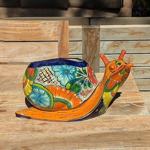 May include: A ceramic snail-shaped planter featuring a vibrant, hand-painted floral pattern in shades of orange, blue, green, yellow, and red. The snail has a glossy finish, a wide opening on its shell, and two prominent antennae on its head.