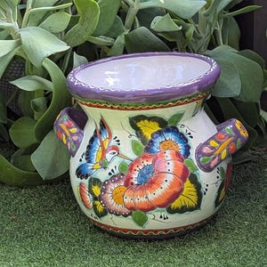 May include: A hand-painted ceramic planter with a wide opening and two handles. The planter features a white base with colorful floral and hummingbird designs in orange, blue, yellow, and green. The rim is purple, and the handles have a decorative pattern.