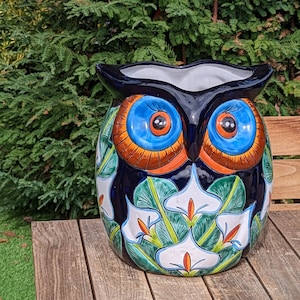 May include: A ceramic owl-shaped planter with a dark blue base, featuring large blue eyes with orange accents. The planter is decorated with white calla lilies and green leaves. It sits on a wooden table outdoors.