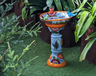 May include: A decorative bird bath with a blue, orange, and white floral design. The bird bath has a pedestal base with a butterfly motif and is topped with three small bird figurines. The bird bath is placed on a green surface, surrounded by plants.