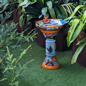May include: A decorative bird bath with a blue, orange, and white floral design. The bird bath has a pedestal base with a butterfly motif and is topped with three small bird figurines. The bird bath is placed on a green surface, surrounded by plants.