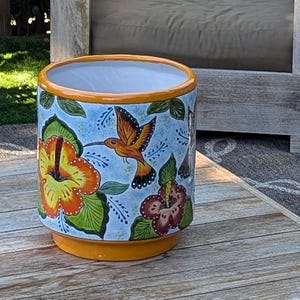 May include: A cylindrical ceramic planter with a vibrant floral and hummingbird design. The planter features a light blue background with orange, yellow, and red hibiscus flowers and a hummingbird. The rim and base are painted orange.