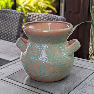 May include: A decorative ceramic vase with two handles. The vase features a peach-colored base with turquoise floral patterns. The vase is sitting on a gray wooden table, with a blurred background of outdoor furniture and greenery.