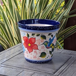May include: A ceramic flower pot featuring a white base with hand-painted designs of a large red flower and a colorful hummingbird. The rim and base are decorated with a dark blue band containing white line details and small dots.