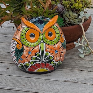 Puede incluir: Figura de búho de cerámica colorida con detalles en naranja, verde y azul. El búho tiene ojos grandes y redondos y está decorado con patrones florales. La figura está sobre una superficie de madera, con una planta en maceta al fondo.