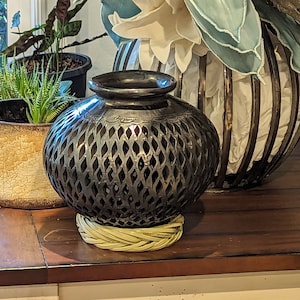 May include: A black ceramic vase with a decorative lattice pattern sits on a wooden table. The vase is on a woven straw base.