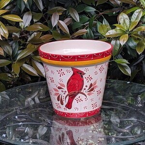 May include: White ceramic planter with a red cardinal bird design. The planter has a red and yellow rim and a white background with red and yellow floral accents.
