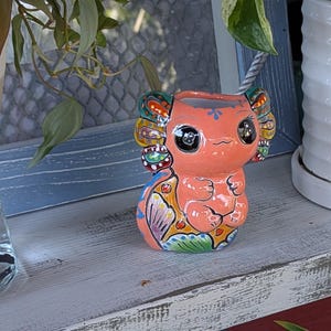 May include: A ceramic planter in the shape of a cartoon creature, primarily orange with colorful accents. The planter has large, expressive eyes and decorative ear-like features. The design includes floral and geometric patterns.
