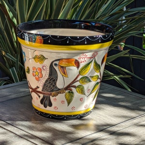 May include: A white ceramic planter with a black rim and a yellow stripe. The planter is decorated with a toucan, leaves, and flowers in shades of yellow, green, orange, and red.