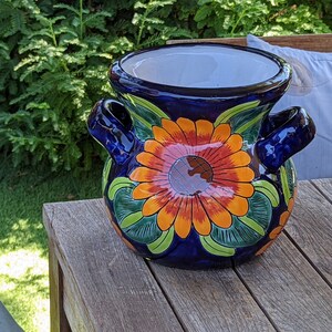 May include: A blue ceramic planter with a floral design. The planter has two handles and features a large orange flower with green leaves.