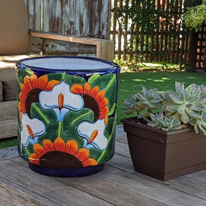 May include: A blue ceramic planter with a colourful floral design featuring orange, white, and green flowers. The planter is sitting on a wooden table.