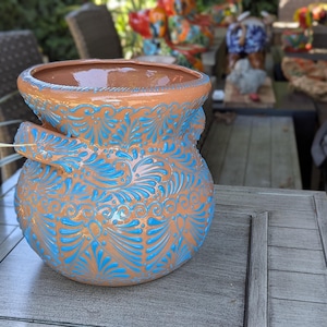 May include: A terracotta clay pot featuring a wide, rounded body and a flared rim. The exterior is decorated with intricate, hand-painted bright blue swirling patterns and leaf-like motifs that cover the entire surface of the vessel.