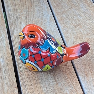 Puede incluir: Una pequeña figura de pájaro de cerámica con un acabado brillante y un patrón multicolor vibrante. El pájaro tiene la cabeza naranja, el pico amarillo y el cuerpo decorado con formas geométricas y motivos florales rojos, azules, verdes y amarillos.