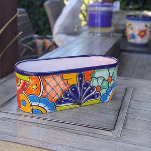 May include: A hand-painted, oval ceramic planter with a vibrant design. The planter has a white interior, a blue rim, and a colourful exterior with floral and geometric patterns in orange, yellow, blue, and green. It sits on a grey wooden surface.