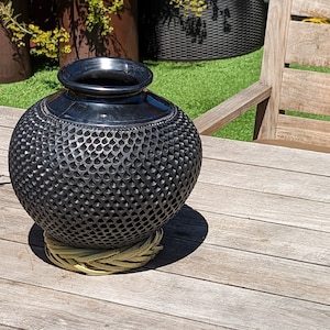 May include: A black, round decorative vase with a textured, patterned surface. The vase sits on a woven, light-yellow coaster. The vase has a wide opening and a smooth, shiny finish. The vase is displayed on a wooden table.