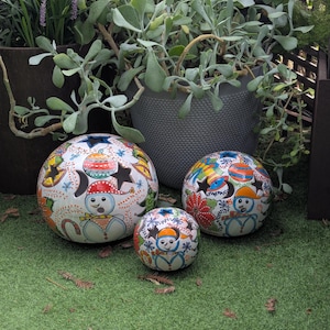 May include: Three spherical ceramic ornaments of varying sizes, each featuring a white base with hand-painted colorful designs of snowmen, stars, moons, and floral patterns. The surfaces include cut-out star shapes and intricate festive details.