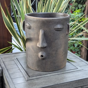 May include: A gray ceramic planter with a face design. The planter has closed eyes, a nose, and a mouth. The planter is sitting on a gray table. Green and yellow plants are in the background.