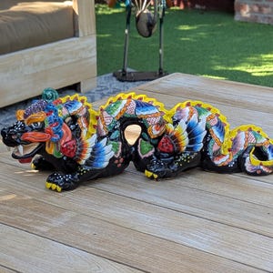 May include: A long, horizontal ceramic dragon figurine featuring a glossy black base color with intricate, multi-colored hand-painted patterns in red, blue, green, and yellow. The dragon has a prominent yellow crest, wings, and sharp teeth.