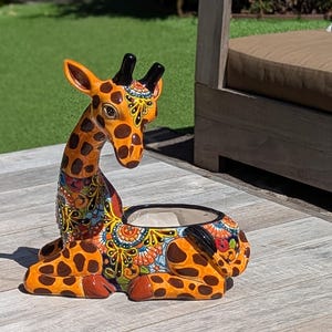 May include: A ceramic giraffe planter with a detailed, colorful design. The giraffe has orange and brown spots, with a decorative pattern around the planter's base. The planter is sitting on a wooden surface.