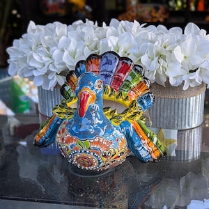 May include: A colorful ceramic turkey figurine with intricate details and a blue, yellow, red, and green paint scheme. The figurine is sitting on a table with a white flower arrangement in the background.