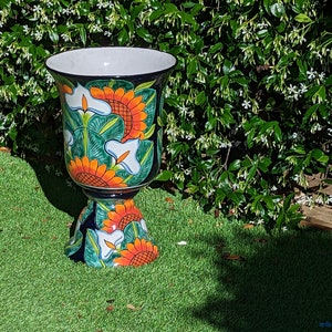 May include: A large, colorful ceramic planter with a pedestal base. The planter features a floral design with sunflowers, white flowers, and green leaves on a blue background.