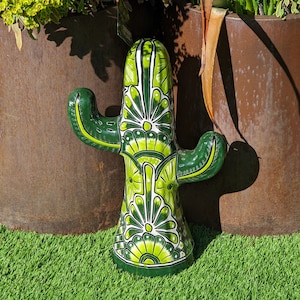 Puede incluir: Una figurita de cactus de cerámica verde y blanca con un diseño floral detallado. El cactus está de pie sobre una superficie de césped artificial verde.