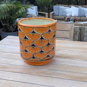 May include: An orange ceramic planter with a repeating pattern of stylized fan shapes in navy blue, white, and orange. The planter has a cylindrical shape and a light green interior. It sits on a wooden table outdoors.